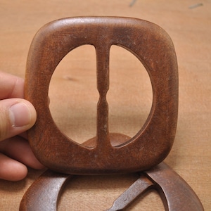 May include: A dark brown wooden buckle with a square shape and a rounded edge. The buckle has two oval-shaped holes in the center.