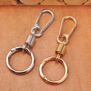 Può includere: Due portachiavi in metallo, uno color argento e uno color oro. Ogni portachiavi è dotato di un moschettone a molla collegato tramite una molla elicoidale cilindrica a un anello circolare, progettato per contenere chiavi o piccoli oggetti.