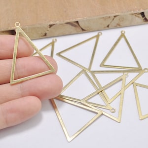 May include: A collection of brass-colored triangle charms with small loops at the top for jewelry making. The charms are open-framed, with clean lines and a matte finish. Several are scattered on a white surface.