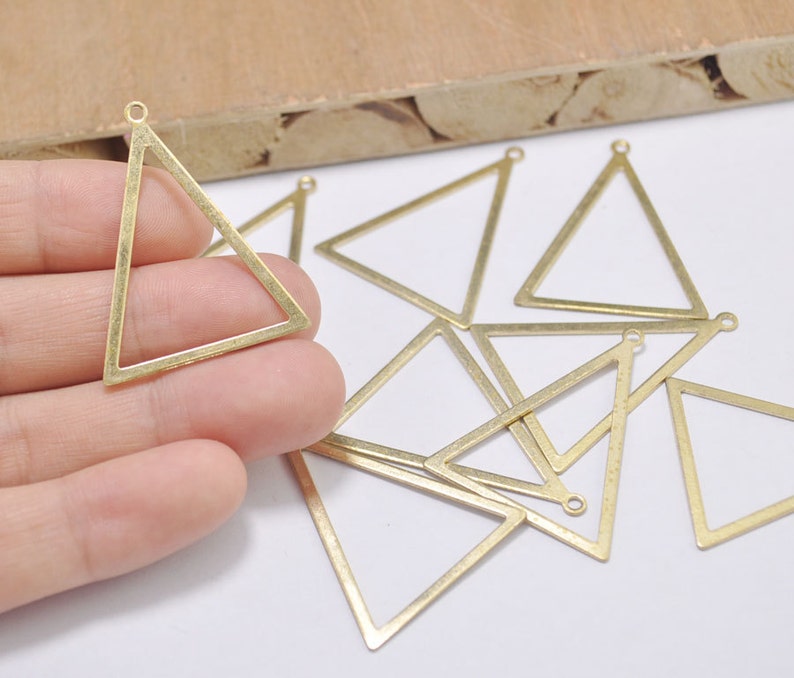 May include: A collection of brass-colored triangle charms with small loops at the top for jewelry making. The charms are open-framed, with clean lines and a matte finish. Several are scattered on a white surface.