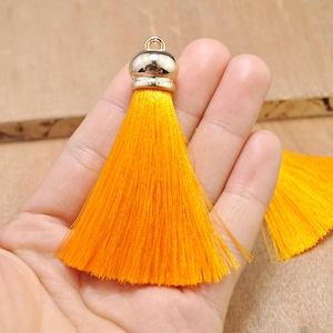 May include: A bright orange tassel with a gold metal top. The tassel is made of soft, silky threads and is approximately 2 inches long.