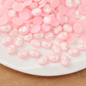 May include: A collection of pink oval and round cabochons with white cameo designs. The cabochons are scattered on a white plate, creating a delicate and feminine aesthetic. Ideal for jewelry making or craft projects.