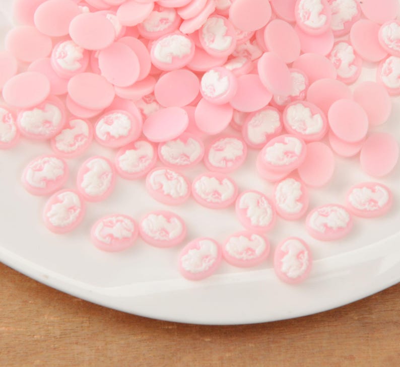 May include: A collection of pink oval and round cabochons with white cameo designs. The cabochons are scattered on a white plate, creating a delicate and feminine aesthetic. Ideal for jewelry making or craft projects.