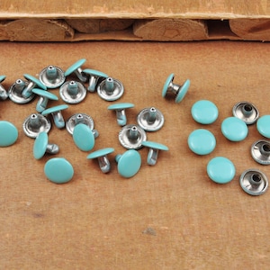 May include: A collection of 20 turquoise metal rivets with silver accents. The rivets are arranged in two groups, one with the rivets in a circular pattern and the other with the rivets in a more random pattern.