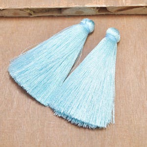 May include: Two light blue silk tassels with a long fringe. The tassels are on a wooden surface.