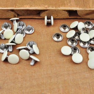 May include: A collection of silver metal and white plastic rivets. The rivets are arranged in two groups on a wooden surface.