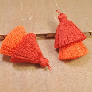 Puede incluir: Dos borlas de adorno de color rojo y naranja con un diseño en capas. Las borlas están hechas de fibras suaves y esponjosas y tienen un aspecto delicado y hecho a mano.