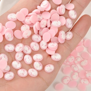 May include: A collection of small, pink cameo cabochons with white relief details. The oval-shaped cabochons feature a classical profile design. These craft supplies are scattered across a surface.