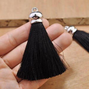 May include: Two black silk tassels with silver metal caps. The tassels are approximately 2 inches long and are held in a hand.