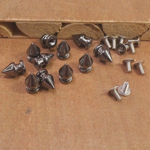 May include: A collection of silver-toned metal pyramid-shaped studs with screws. The studs have a pointed top and a flat base. The screws are silver-toned and have a Phillips head.