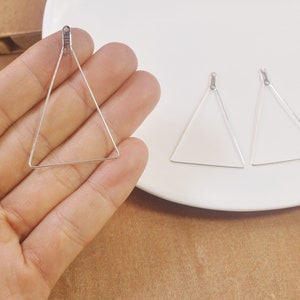 May include: Three silver triangle earring blanks with jump rings. The earring blanks are on a white plate and a hand is holding one of the blanks.