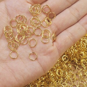 Puede incluir: Primer plano de una mano sosteniendo una gran cantidad de anillos de salto dorados. Los anillos son peque&ntilde;os y delicados, y son perfectos para la elaboraci&oacute;n de joyas.