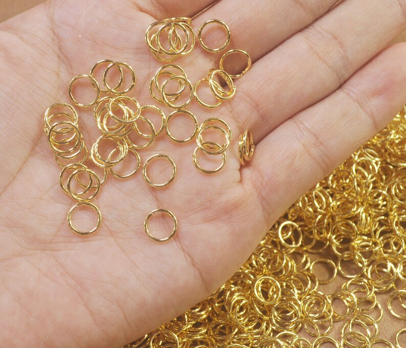 Puede incluir: Primer plano de una mano sosteniendo una gran cantidad de anillos de salto dorados. Los anillos son peque&ntilde;os y delicados, y son perfectos para la elaboraci&oacute;n de joyas.