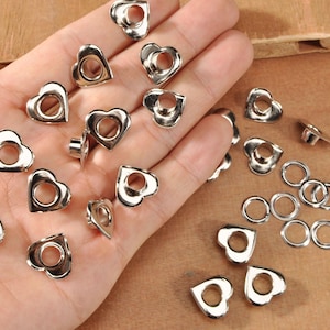 May include: A collection of silver-colored heart-shaped eyelets and small round eyelets. The eyelets are scattered on a light brown surface, with some held in a hand. The heart-shaped eyelets have a hole in the center.