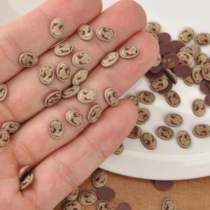 May include: A collection of small, oval-shaped polymer clay beads. The beads are light tan with a darker brown design on the surface. Some dark brown oval beads are also present. The beads are scattered on a white surface and held in a hand.