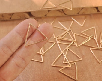 May include: A close-up of a hand holding a gold triangle pendant. The pendant is made of thin wire and has a simple, geometric design. There are many other gold triangle pendants scattered on a wooden surface in the background.