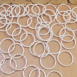 May include: A pile of silver jump rings, each with a twisted, textured design. The rings are scattered on a light brown wooden surface.