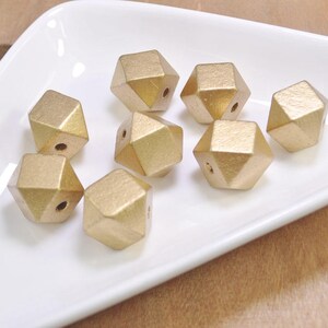 Może przedstawiać: Grupa ośmiu złotych drewnianych koralików, każdy o geometrycznym, fasetowanym kształcie. Korale są ułożone na białej powierzchni.