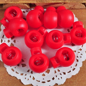 Puede incluir: Primer plano de diez cierres de palanca de plástico rojo con un resorte de metal. Los cierres están dispuestos en círculo sobre una servilleta blanca.