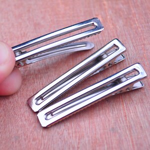 Op de afbeelding: Close-up van verschillende zilveren metalen haarclips. De clips zijn rechthoekig met een scharnierend ontwerp en een geribbeld interieur voor grip. De clips worden weergegeven op een lichtbruin houten oppervlak.
