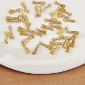 Puede incluir: Una colección de pequeñas borlas doradas con detalles plateados y aros metálicos. Las borlas están dispuestas sobre una superficie blanca y ligeramente curvada, sugiriendo un tema de artesanía o joyería.