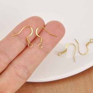 May include: Close-up of several gold-colored earring hooks. Two hooks are held in a hand, while two more are on a white surface. The hooks have a curved design with a coil and a small loop at the bottom.