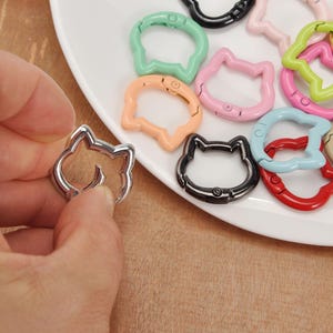 Puede incluir: Una mano sostiene un cierre plateado con forma de gato. Otros cierres con forma de gato en varios colores, como verde, rosa y negro, est&aacute;n dispuestos sobre un plato blanco. Los cierres son de metal y tienen un mecanismo de resorte.