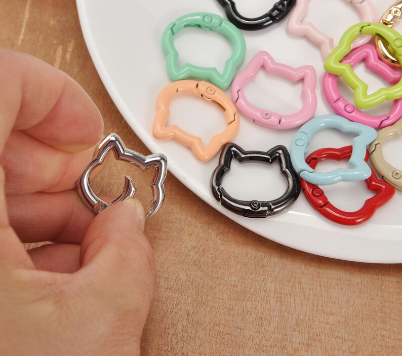 Puede incluir: Una mano sostiene un cierre plateado con forma de gato. Otros cierres con forma de gato en varios colores, como verde, rosa y negro, est&aacute;n dispuestos sobre un plato blanco. Los cierres son de metal y tienen un mecanismo de resorte.