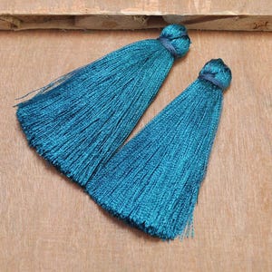 May include: Two teal blue silk tassels with a knotted top. The tassels are on a wooden surface.
