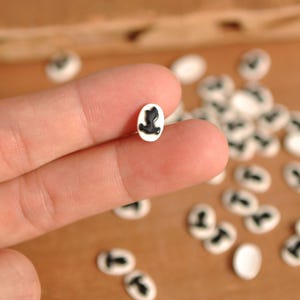May include: A collection of small, white oval-shaped beads with a black silhouette design. One bead is held between fingers, showcasing the detail. The beads are scattered on a light brown surface.
