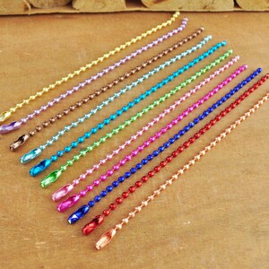 May include: Assortment of 14 colourful metal chain links, each with a different colour, including gold, silver, purple, brown, blue, green, pink, red, and copper. The chains are laid out in a row on a wooden surface.