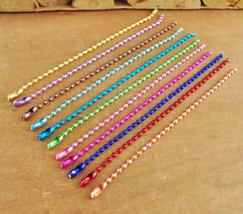 May include: Assortment of 14 colourful metal chain links, each with a different colour, including gold, silver, purple, brown, blue, green, pink, red, and copper. The chains are laid out in a row on a wooden surface.