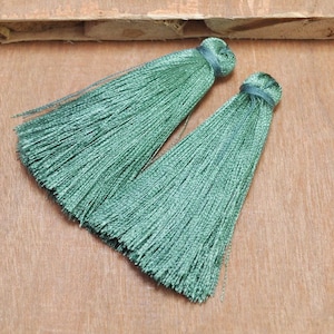May include: Two sage green tassels with a silky texture. Each tassel is bound at the top with a darker green thread. The tassels are lying on a light brown wooden surface.