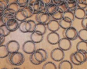 May include: A scattering of small, textured metal rings on a wooden surface. The rings are a dark, metallic colour and feature a detailed, etched pattern around their circumference. The image is a top-down view, showing the quantity and arrangement of the rings.