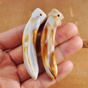 May include: Two white and brown ceramic tooth-shaped pendants with a hole at the top for hanging.