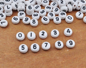 Può includere: Perline rotonde bianche con numeri neri da 0 a 9. Le perline sono sparse su una superficie di legno. Alcune perline sono cilindriche, altre piatte. Ideali per la creazione di gioielli o l'artigianato.