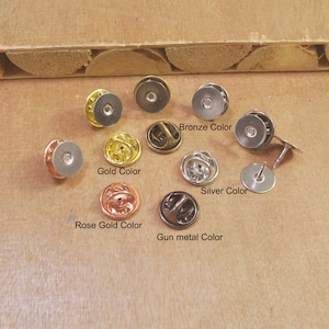 Puede incluir: Surtido de 10 respaldos de pines metálicos en diferentes colores, incluyendo bronce, oro, oro rosa, plata y metal gris. Los respaldos de pines están dispuestos en un círculo sobre una superficie de madera.