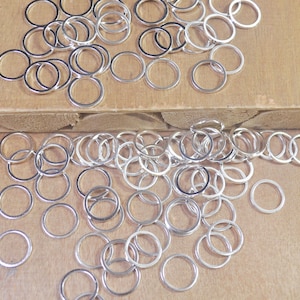 May include: A large pile of silver jump rings, a type of jewelry finding, scattered on a wooden surface. The rings are all the same size and are made of a shiny metal.