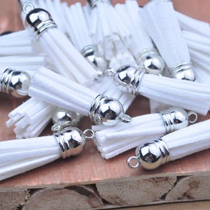 Peut inclure: Plusieurs pompons blancs avec des capuchons et des boucles en métal argenté. Les pompons sont faits d'un matériau doux et texturé, disposés sur une surface en bois. Idéal pour la création de bijoux ou les projets d'artisanat.