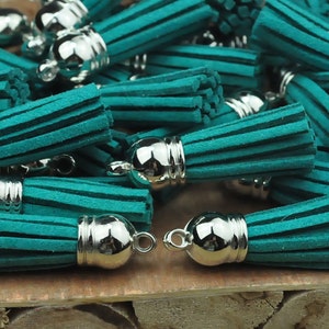May include: A group of teal green suede tassels with silver metal caps. The tassels are arranged in a pile on a wooden surface.