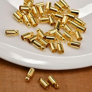 May include: A collection of gold-toned metal cord end caps, each featuring a cylindrical body with a small loop at the top. These jewelry findings are scattered across a white surface, showcasing a polished, reflective finish and uniform design.