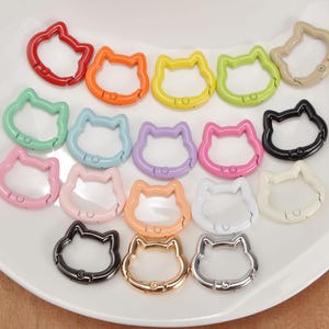 Puede incluir: Una colecci&oacute;n de mosquetones con forma de gato de colores, incluyendo rojo, naranja, amarillo, verde, azul, morado, rosa, negro, blanco y dorado. Estos clips met&aacute;licos est&aacute;n dispuestos sobre una superficie blanca.