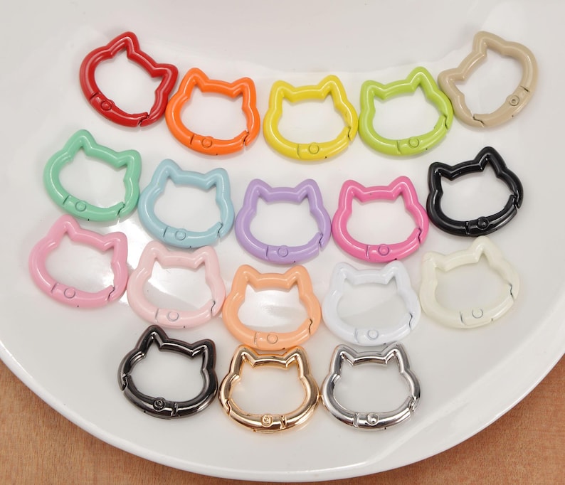 Puede incluir: Una colecci&oacute;n de mosquetones con forma de gato de colores, incluyendo rojo, naranja, amarillo, verde, azul, morado, rosa, negro, blanco y dorado. Estos clips met&aacute;licos est&aacute;n dispuestos sobre una superficie blanca.