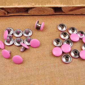 May include: A collection of pink metal rivets with a silver finish. The rivets are arranged in a circle on a wooden surface.