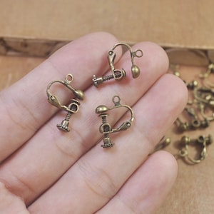 Peut inclure: Gros plan de plusieurs apprêts de boucles d'oreilles à clip de couleur bronze antique. Les apprêts ont une conception à charnière avec un mécanisme à vis pour régler la tension. Les boucles d'oreilles sont petites et délicates, avec une esthétique vintage.