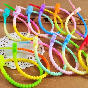 Puede incluir: Un conjunto de bridas de cable de silicona multicolores, arcoíris. Las bridas están hechas de un material flexible y plegable y tienen un pequeño botón redondo de plástico en el extremo.