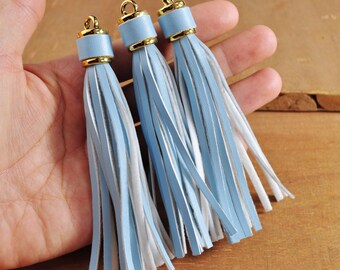 May include: Three light blue leather tassels with gold metal tops. The tassels are hanging from a hand.