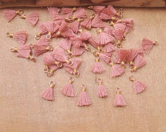 May include: A collection of pink tassel charms with gold accents. The tassels are scattered on a wooden surface.
