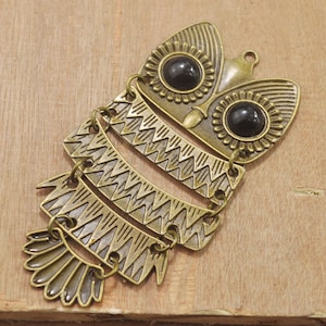 Könnte beinhalten: Ein goldfarbener Metallamulet in Form einer Eule mit schwarzen Glasaugen. Die Eule ist mit einem geschichteten Design verziert und hat einen baumelnden federartigen Schwanz.