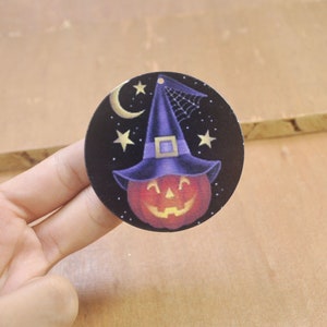 May include: A round, black and white Halloween ornament with a pumpkin wearing a purple witch hat and a crescent moon with stars in the background.